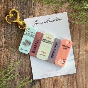 May include: A keychain with miniature book charms, including titles such as "Pride & Prejudice" and "Emma" by Jane Austen. The charms are in pastel shades and attached to a gold-coloured heart-shaped clasp.