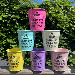 May include: Six brightly coloured metal buckets stacked together. Each bucket is a different colour: pink, turquoise, cream, yellow, lilac, and pale pink. The text "thank you for helping me grow" is printed in black on each bucket.