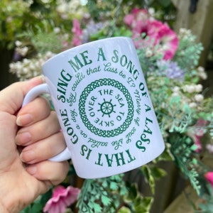 May include: White ceramic mug with a green design that says "That is gone sing me a song of a lass" with a Celtic knot border and a graphic of a ship's wheel with the words "The Sea to Skye Over" around it.