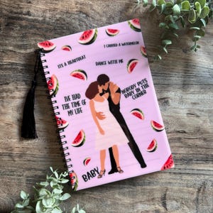 May include: A pink spiral-bound notebook with a cover design featuring a dancing couple, watermelon slices, and phrases such as "I carried a watermelon" and "Nobody puts Baby in the corner." It has a black tassel.