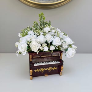 May include: A decorative floral arrangement in the shape of a miniature piano. The dark brown piano has gold detailing and a keyboard. The top is filled with white roses and other white flowers and greenery, creating a decorative centerpiece.