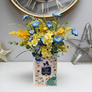 May include: A decorative vase filled with yellow and blue flowers. The vase is designed to resemble a book, featuring the title "Pride and Prejudice" and a peacock design. The flowers are a mix of sizes and shapes, creating a vibrant display.