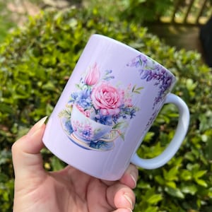May include: A lavender mug with a floral design featuring a teacup filled with pink and purple flowers. The design includes a saucer with a floral pattern.