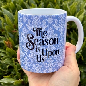 Puede incluir: Una taza de cer&aacute;mica blanca con un patr&oacute;n de damasco azul y el texto "The Season is Upon Us" en negro. La taza tiene un asa blanca y est&aacute; sujeta con una mano.