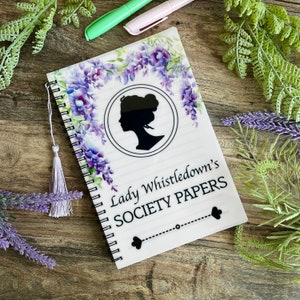May include: A white spiral bound notebook with a purple floral design and a silhouette of a woman's head. The notebook has the text "Lady Whistledown's Society Papers" printed on the front.