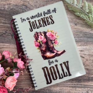 May include: A notebook with a spiral binding and a decorative cover. The cover displays the text "In a world full of Jolenes, Be a Dolly" above an illustration of brown cowboy boots with pink roses. A burgundy tassel hangs from the spiral binding.