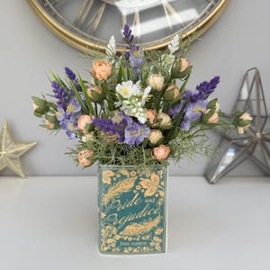 May include: A decorative vase filled with flowers. The vase is teal with gold detailing and the words "Pride and Prejudice" and "Jane Austen". The flowers are a mix of lavender, peach, white, and green, creating a colourful display.