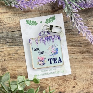 May include: A square keychain with a floral tea-themed design. The keychain has a light blue background with purple wisteria, a teapot, and the words "I am the TEA." It has a silver clasp for easy attachment.
