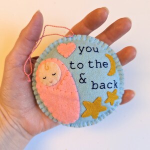 K&ouml;nnte beinhalten: Ein hellblauer Filz-Anh&auml;nger mit einem rosa Filz-Baby, einem rosa Herz, einem goldenen Halbmond und zwei goldenen Sternen. Der Anh&auml;nger ist mit dem Text "you to the & back" bestickt.