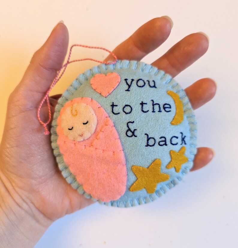 K&ouml;nnte beinhalten: Ein hellblauer Filz-Anh&auml;nger mit einem rosa Filz-Baby, einem rosa Herz, einem goldenen Halbmond und zwei goldenen Sternen. Der Anh&auml;nger ist mit dem Text "you to the & back" bestickt.