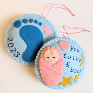 K&ouml;nnte beinhalten: Zwei Filz-Anh&auml;nger, einer blau mit einem Fu&szlig;abdruck und dem Jahr 2023, der andere hellblau mit einem rosa gewickelten Baby, zwei goldenen Sternen, einem rosa Herzen und dem Text "you to the & back".