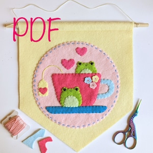 May include: A PDF pattern for a felt wall hanging featuring two green frogs sitting in a pink teacup with a teabag and hearts. The teacup is on a blue saucer. The pattern is on a yellow background.