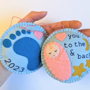 K&ouml;nnte beinhalten: Zwei Filz-Anh&auml;nger, einer blau mit einem Baby-Fu&szlig;abdruck und dem Jahr 2023, der andere hellblau mit einem rosa gewickelten Baby, einem Herz, Sternen und dem Text "you to the & back"