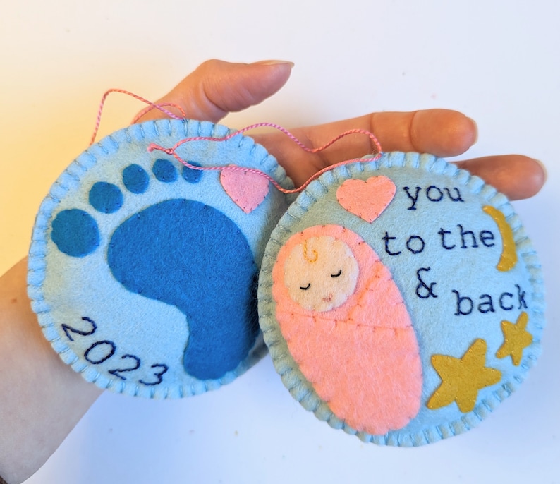 K&ouml;nnte beinhalten: Zwei Filz-Anh&auml;nger, einer blau mit einem Baby-Fu&szlig;abdruck und dem Jahr 2023, der andere hellblau mit einem rosa gewickelten Baby, einem Herz, Sternen und dem Text "you to the & back"