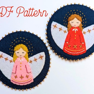 May include: Two felt angel decorations with embroidered details. The angels have blonde hair, white wings, and are wearing dresses. One angel is wearing a red dress with gold stars, and the other is wearing a pink dress with gold stars. The decorations are on a white background.