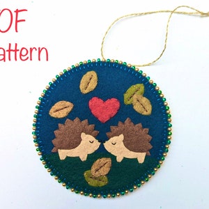 May include: A felt ornament featuring two brown hedgehogs kissing with a red heart above them. The ornament is surrounded by green leaves and gold beads.