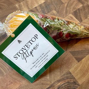 Puede incluir: Una etiqueta verde y blanca con el texto "Stovetop Potpourri" e instrucciones de uso. La etiqueta está unida a una bolsa de plástico transparente llena de rodajas de naranja, arándanos rojos y ramitas de romero.