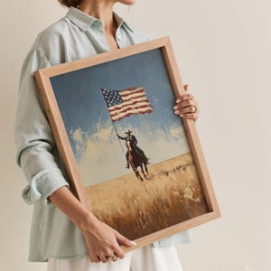 May include: A framed painting depicts a cowboy on horseback waving an American flag against a blue sky and golden field. The artwork is in a wooden frame.