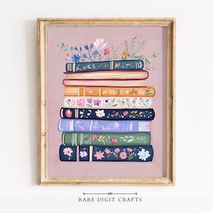 Puede incluir: Una impresión artística enmarcada que presenta una pila de libros con diseños florales y lomos coloridos. Los libros están adornados con varias flores y vegetación, sobre un fondo rosa suave. El marco es de color dorado cálido. El texto "RARE DIGIT CRAFTS" está en la parte inferior.