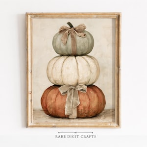 May include: A framed art print featuring a vertical stack of three pumpkins in muted tones. The top pumpkin is sage green, the middle is cream, and the bottom is burnt orange. Each pumpkin is adorned with a tied fabric ribbon bow in a neutral colour.