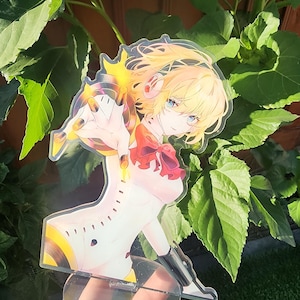 May include: A clear acrylic anime figure of a blonde character with blue eyes, wearing a white outfit with red accents. The figure is supported by a clear, circular base and is set against a backdrop of green leaves.