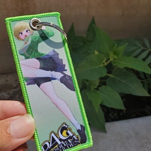 May include: Green and white keychain featuring a cartoon anime character wearing a green skirt and white socks. The keychain has the text "P4G Persona 4 Golden" on it.