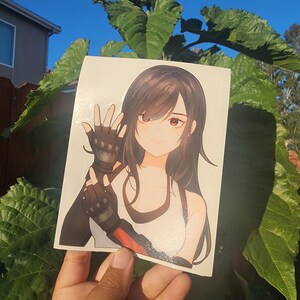 K&ouml;nnte beinhalten: Ein Aufkleber mit einer Anime-Illustration einer Frau mit langen, dunklen Haaren, die ein wei&szlig;es Tanktop mit schwarzem Besatz und fingerlose Handschuhe tr&auml;gt. Der Hintergrund ist ein klarer blauer Himmel und gr&uuml;nes Laub.
