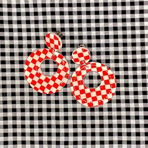 Puede incluir: Un par de pendientes a cuadros rojos y blancos. Cada pendiente tiene un pequeño perno redondo y un aro circular más grande con un agujero en el centro. Los pendientes están sobre un fondo a cuadros blanco y negro.