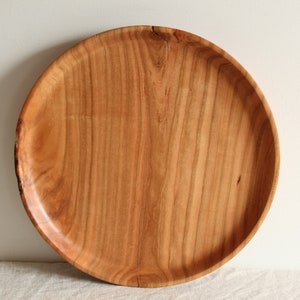 May include: A large, round wooden serving tray with a natural wood finish. The tray has a smooth, polished surface and a slightly raised edge.