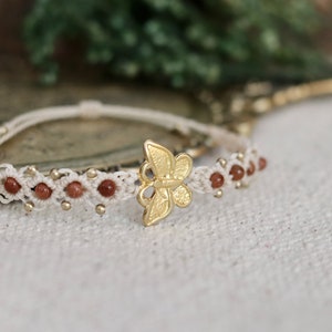 Könnte beinhalten: Ein weißes Makramee-Armband mit braunen Perlen und einem goldenen Schmetterlingsanhänger.