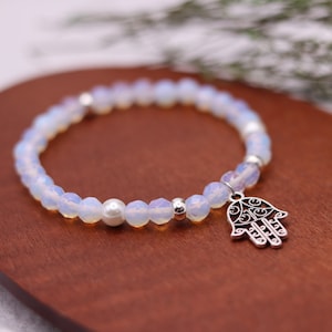 May include: A beaded bracelet featuring iridescent, faceted beads and a silver-tone Hamsa hand charm. The bracelet includes a central pearl bead and silver accent beads. The bracelet is displayed on a wooden surface.
