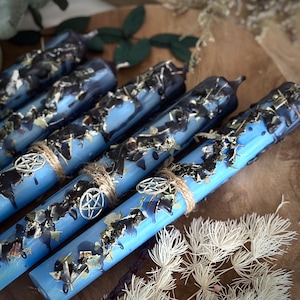 Può includere: Un set di candele blu e nere con un ciondolo a pentagramma e spago. Le candele sono incassate con erbe secche e hanno un aspetto maculato.