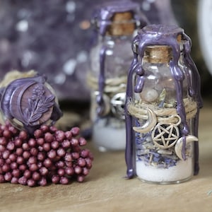 Può includere: Una piccola bottiglia di vetro piena di erbe secche, cristalli e un ciondolo a pentagramma d'argento. La bottiglia è sigillata con un tappo di sughero e sormontata da un sigillo di cera viola. La bottiglia è circondata da uno spago rustico e un ciondolo a mezzaluna d'argento.