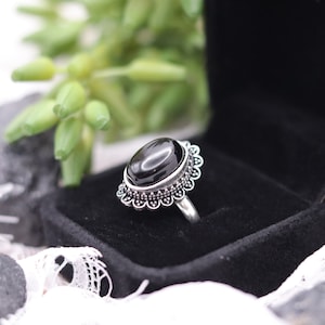 Könnte beinhalten: Ein Silberring mit einem glatten, ovalen schwarzen Stein, der in einer dekorativen, gewellten Metallfassung sitzt. Der Ring besteht aus schlichtem Silber und die Fassung weist um den schwarzen Edelstein herum feine, perlenartige Details auf.