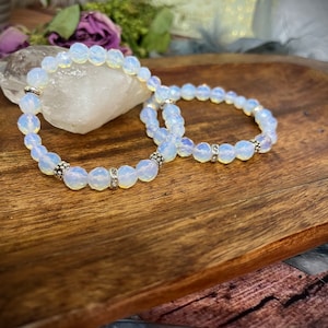 Könnte beinhalten: Zwei weiße Opal-Perlenarmbänder mit silbernen Abstandshaltern. Die Armbänder liegen auf einer Holzoberfläche.