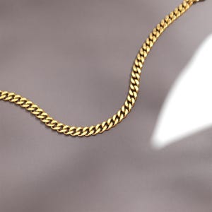 K&ouml;nnte beinhalten: Eine goldene Kettenkette im Curb-Link-Stil. Die Kette liegt flach auf einer grauen Oberfl&auml;che.