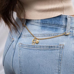 K&ouml;nnte beinhalten: Eine goldene Kette mit einem Anh&auml;nger, der die Zahl "5" und einen Fu&szlig;ball zeigt, h&auml;ngt an der Ges&auml;&szlig;tasche einer hellblauen Jeans.
