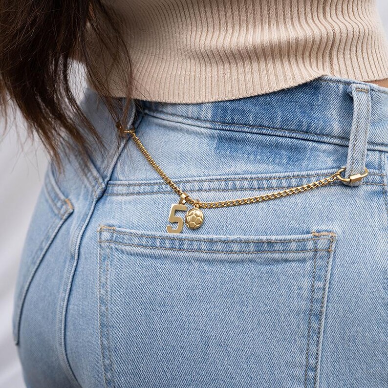 K&ouml;nnte beinhalten: Eine goldene Kette mit einem Anh&auml;nger, der die Zahl "5" und einen Fu&szlig;ball zeigt, h&auml;ngt an der Ges&auml;&szlig;tasche einer hellblauen Jeans.