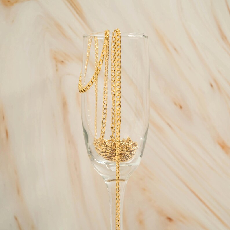 K&ouml;nnte beinhalten: Eine goldene Kettenkette, die in einer klaren Glas-Champagnerfl&ouml;te drapiert ist.