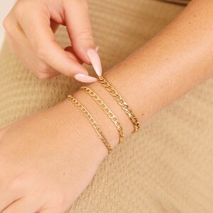 K&ouml;nnte beinhalten: Vier goldene Kettenarmb&auml;nder, die auf dem Handgelenk einer Person &uuml;bereinander liegen. Die Armb&auml;nder haben unterschiedliche Breiten und ein klassisches Curb-Ketten-Design.