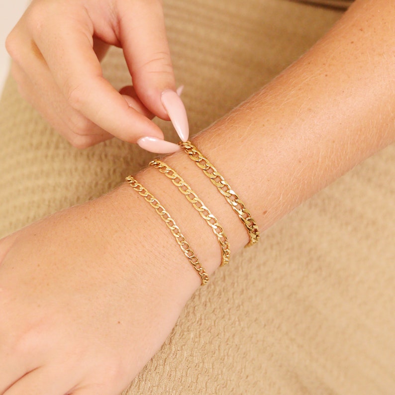 K&ouml;nnte beinhalten: Vier goldene Kettenarmb&auml;nder, die auf dem Handgelenk einer Person &uuml;bereinander liegen. Die Armb&auml;nder haben unterschiedliche Breiten und ein klassisches Curb-Ketten-Design.