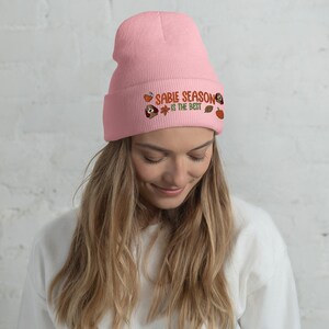 Puede incluir: Un gorro rosa claro con la frase bordada "SABIE SEASON IS THE BEST" en naranja y verde, junto con gráficos de otoño. El gorro está hecho de un material suave y tejido.