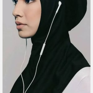 May include: A black athletic hijab with a white cord running through it. The hijab is worn by a person with dark hair and brown eyes. The person is wearing a white shirt underneath the hijab.