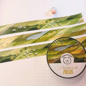 Peut inclure: Rouleau de ruban adhésif washi décoratif avec un motif de paysage de champs verts, d'un lac et de moutons. Le rouleau de ruban adhésif porte le texte "Patchwork Fields" et un logo d'ours. Le ruban est déroulé.
