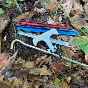May include: A close-up of a silver tent stake with a hook and a black cord. The stake is lying on a bed of brown leaves and green grass.
