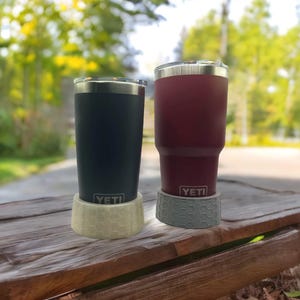 May include: Two YETI brand stainless steel tumblers with lids, one is navy blue and the other is burgundy. Both tumblers are sitting on a wooden surface with a gray and beige patterned coaster underneath each.