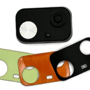 Puede incluir: Dispositivo rectangular negro con un altavoz circular y un círculo plateado con patrón radial. Tres cubiertas rectangulares de colores en verde, naranja y negro con recortes circulares y rectangulares.