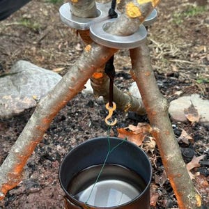 Könnte beinhalten: Ein silberner Dreibeinständer mit einem daran hängenden Metallhaken. Ein Topf mit Wasser hängt am Haken über einem Feuer.