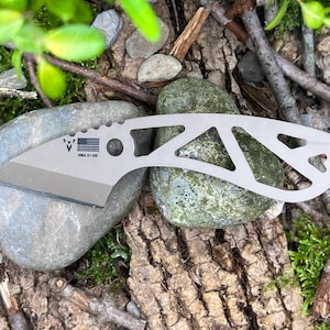 Puede incluir: Un cuchillo de bolsillo plateado con un mango esqueletizado y una hoja parcialmente dentada. El cuchillo tiene un pequeño agujero para un cordón y el texto "KOBRA 6/130" está grabado en la hoja.