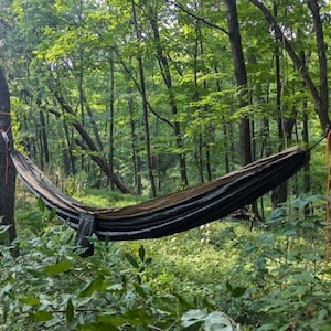 May include: A black hammock is strung between two trees in a woodland. The hammock is made of woven fabric and has a dark green and black pattern. The trees are tall and have green leaves. The hammock is hanging low to the ground.